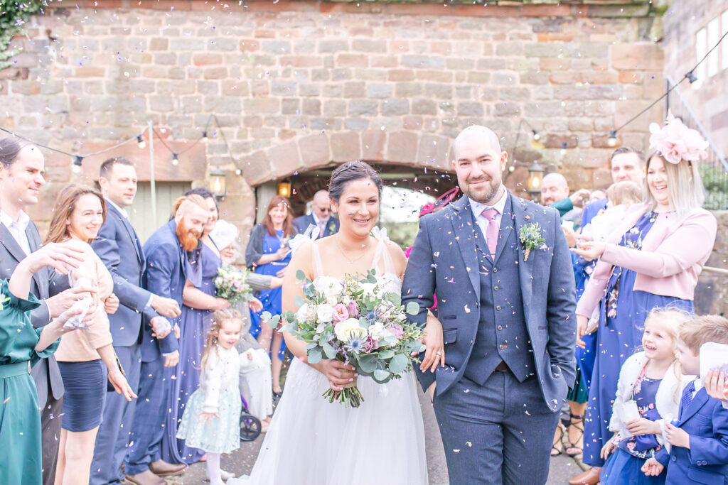 Abby and Jamie arm in arm walking while their guests throw confetti on them in the grounds at The Ashes Wedding Barns in Staffordshire